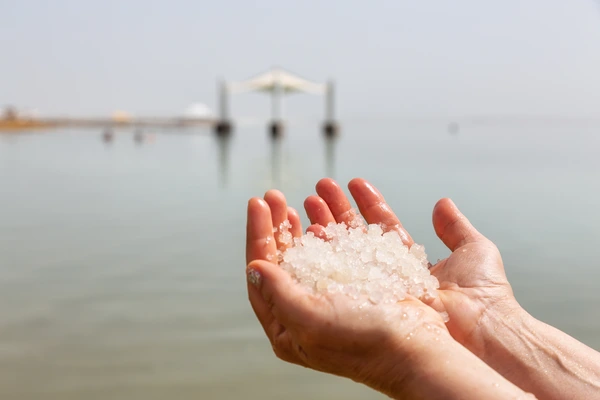 Middle age woman holding salt crystals in her hand with the Dead Sea in the Background during a bright sunny day. Taken in Ein Bokek, Israel.