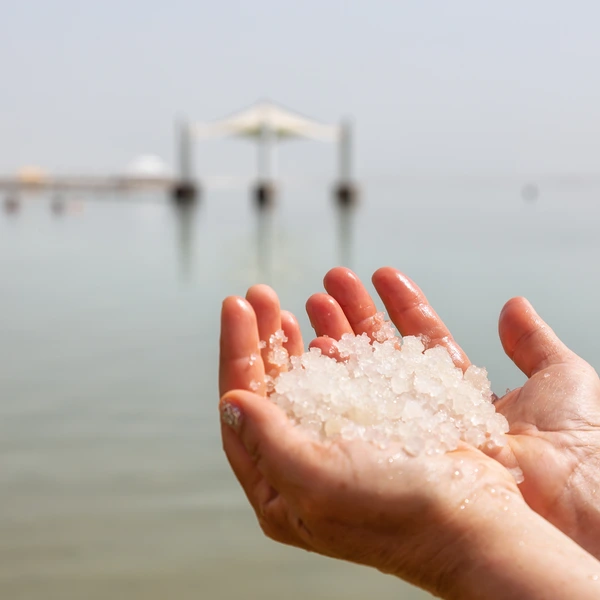 Middle age woman holding salt crystals in her hand with the Dead Sea in the Background during a bright sunny day. Taken in Ein Bokek, Israel.